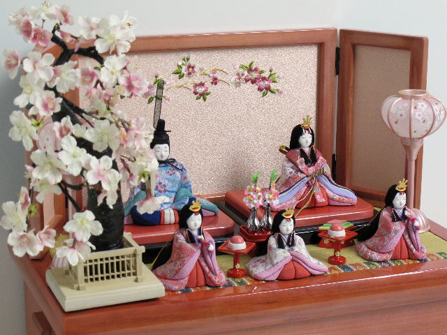 女雛は白とピンク、男雛はブルーの桜花びら柄衣装の木目込み人形五人揃いです。桜の刺繍がポイントのパールローズピンクの収納台に飾りました。