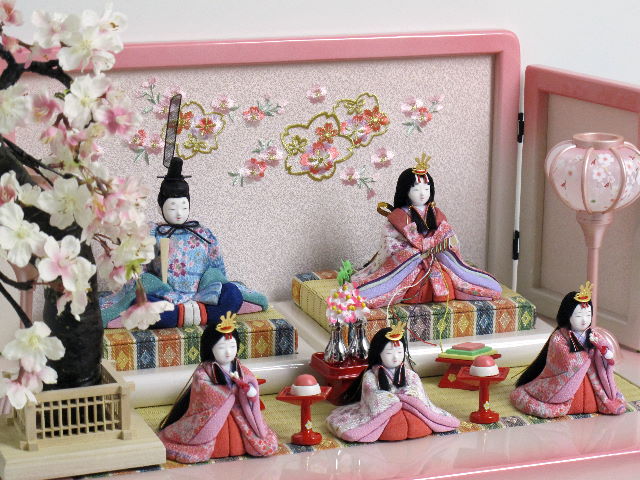 女雛は白とピンク、男雛はブルーの桜花びら柄衣装の木目込み人形五人揃いです。桜柄の刺繍がポイントのホワイトぼかしピンクの収納台に飾りました。