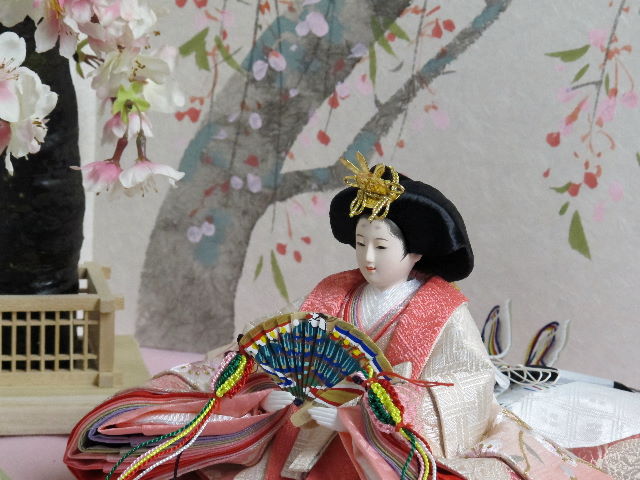 桜の刺繍が特徴の姫はピンク、殿はグリーンのグラデーションが綺麗な雛人形を枝垂桜と桜絵屏風で優雅に飾った桐箱収納式のひな人形です。