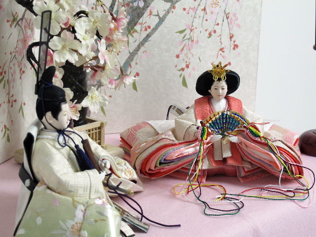 桜の刺繍が特徴の姫はピンク、殿はグリーンのグラデーションが綺麗な雛人形を枝垂桜と桜絵屏風で優雅に飾った桐箱収納式のひな人形です。
