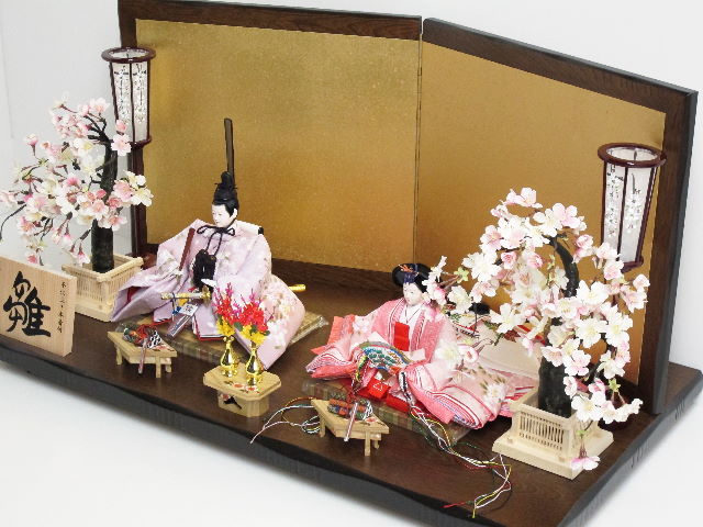 おとなしいピンクの地に桜をデザインした優しい雰囲気の衣装で仕立て自慢のおひなさまを創作金屏風と巻物前飾り、左右の桜で重厚に飾った雛人形です。